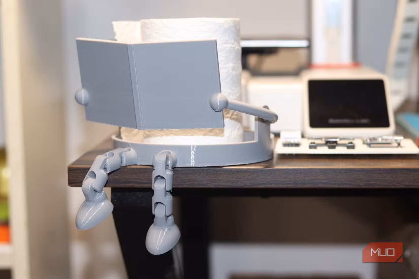Держатель для туалетной бумаги в виде читающего персонажа рядом с 3D-принтером